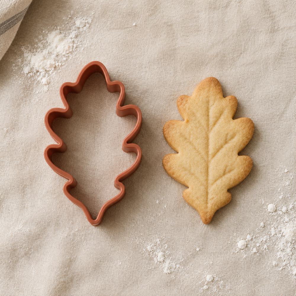Cortador de galletas con forma de hoja junto a una galleta horneada con la misma forma